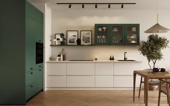 Moderne Küchenzeile in Grün und Weiß mit Glas-Hängeschränken, offener Ablage mit gerahmten Fotos, schwarzem Spülbecken und Armatur; links grüner Hochschrank mit Backofen, rechts Holztisch und Zimmerbaum, Fischgrät-Parkett und Deckenstrahler.